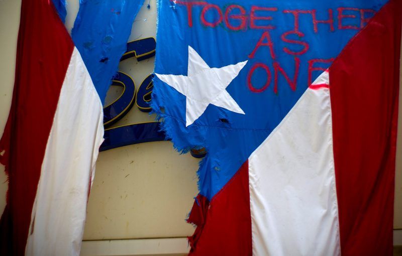 Una dañada bandera puertorriqueña, sobre la que han pintado en aerosol la frase "juntos como si fuéramos uno", cuelga sobre la fachada de un negocio en San Juan, Puerto Rico, el 27 de septiembre de 2017. Credit Ramon Espinosa/Associated Press