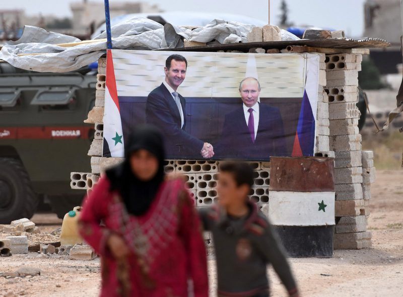 A Syrian woman walks with a boy past a banner showing Russian President Vladimir Putin (R) shaking hands with Syrian President Bashar al-Assad, after arriving in a convoy carrying displaced people into government-controlled territory at Abu al-Zuhur checkpoint in the western countryside of Idlib province, on June 1, 2018. (Photo by George OURFALIAN / AFP) (Photo credit should read GEORGE OURFALIAN/AFP/Getty Images)